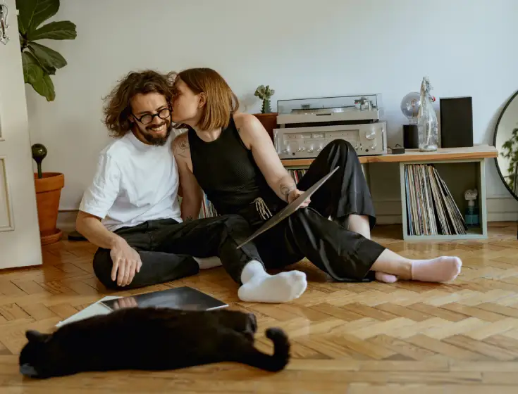 A white young adult couple sit on the wooden floor in their home. The woman is leaning over to kiss her male partner’s cheek, while she holds a record sleeve in one hand. Their black cat is lounging in front of them. 