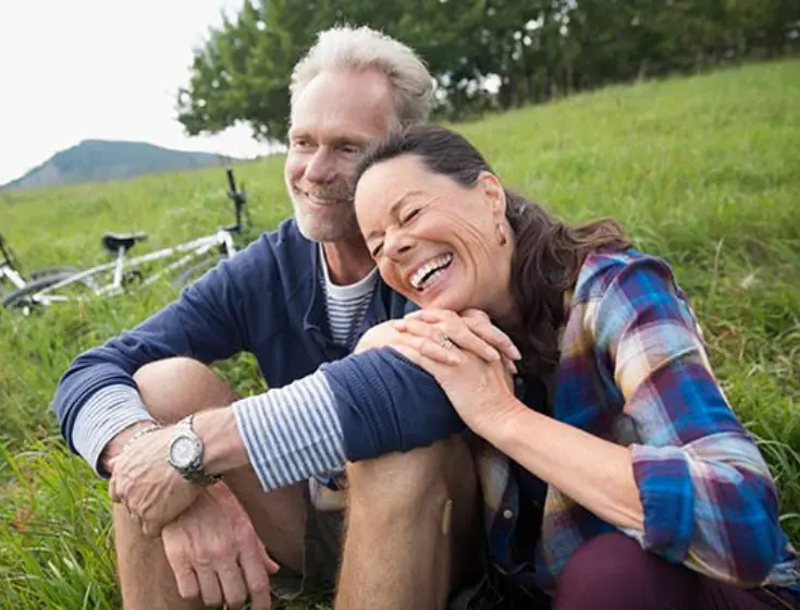 An older couple sit on a grassy hillside together. A bicycle lies in the grass nearby. The man smiles and looks into the distance, while his wife leans against his shoulder laughing. 
