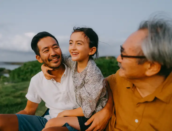 An Asian father holds his young daughter on his lap. Her arm is loosely wrapped around his neck and shoulders. Her grandfather sits beside her and smiles at them. They’re sitting outside near a lake. 