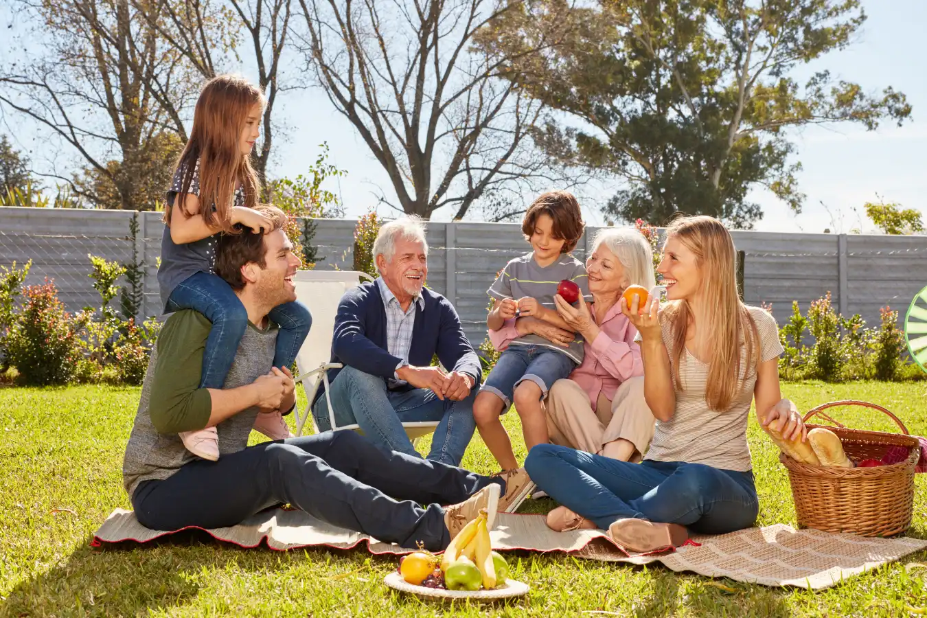 A multigenerational white family are sitting outside on their patio in the sun. One young child sits on their father’s shoulders, while another is held by their grandmother. 