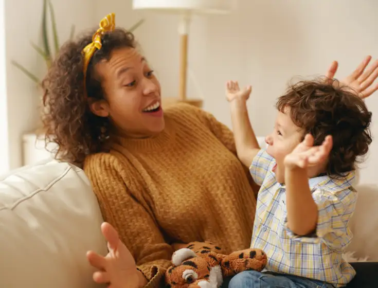 A young olive-skinned woman with short curly hair and yellow headband plays with her young toddler. He has his hands thrown up in the air in playful surprise. His mother mimics the gesture. 