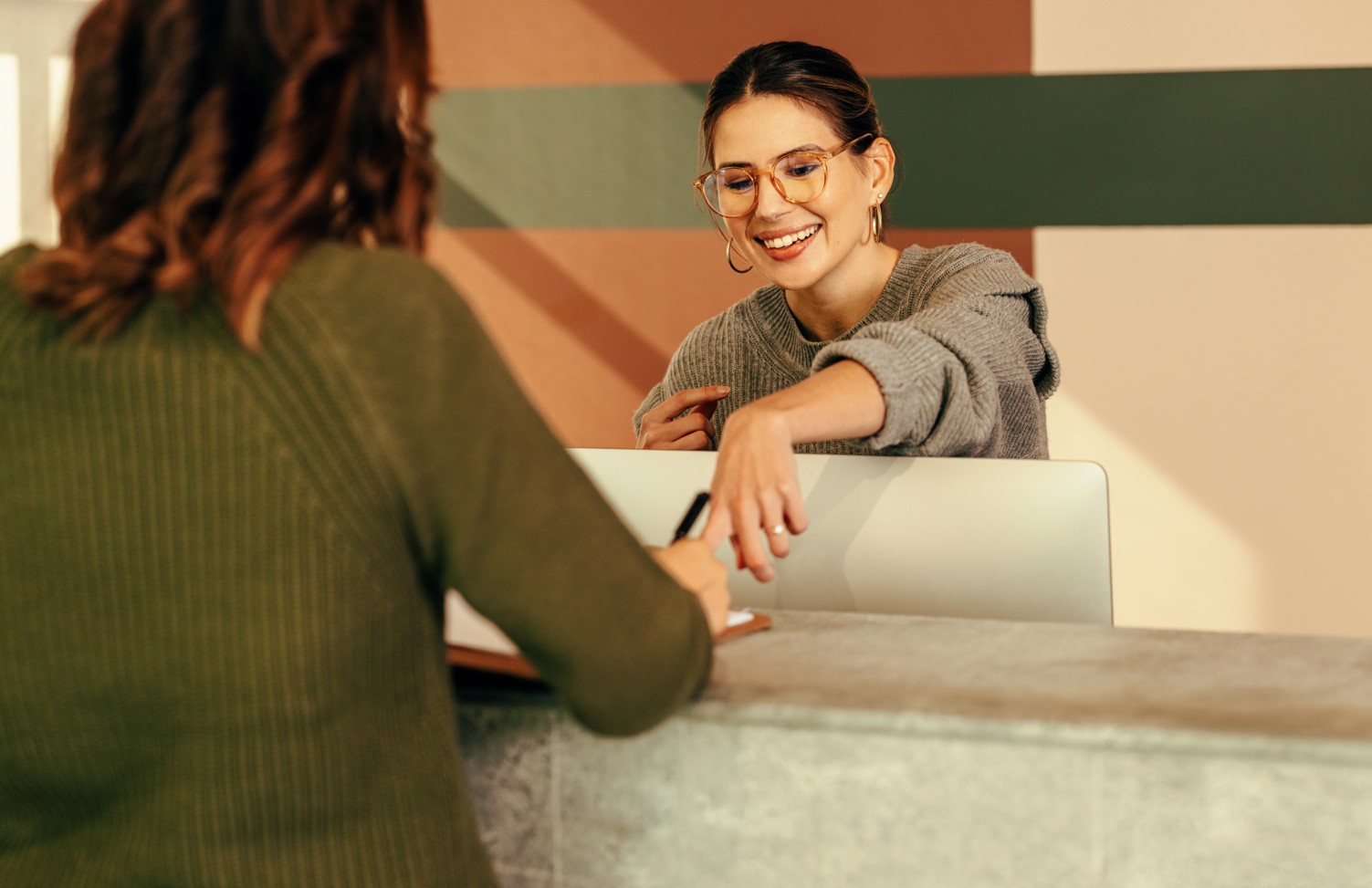 Reception desk interaction with one person writing on a clipboard and another person behind the desk near a monitor.