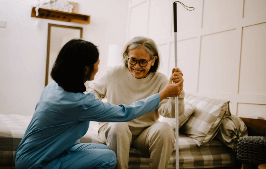A female doctor faces away from the camera as she talks with a senior Asian woman. The senior woman is wearing glasses and a hospital gown. She is smiling as she and the doctor steady a nearby IV stand.