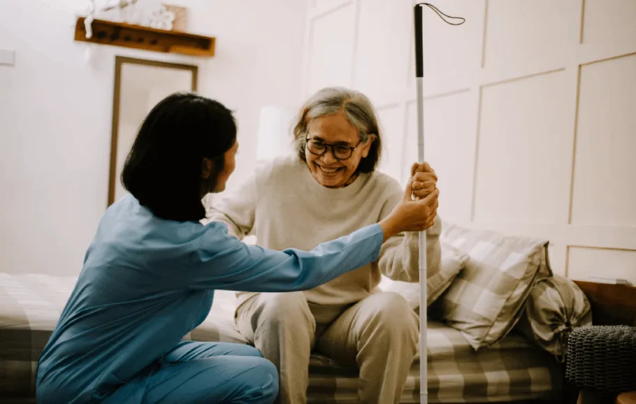 A female doctor faces away from the camera as she talks with a senior Asian woman. The senior woman is wearing glasses and a hospital gown. She is smiling as she and the doctor steady a nearby IV stand. 