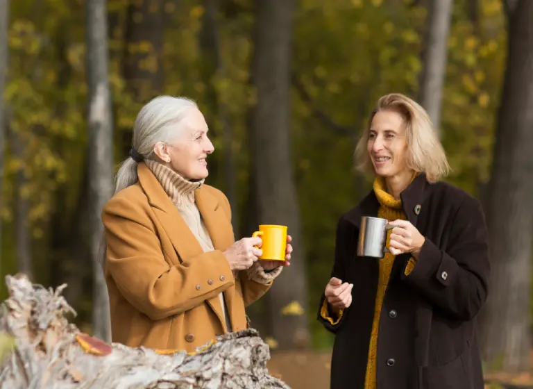 A senior white woman shares a cup of tea with her adult daughter outside. They are both dressed warmly as they stand in a backyard with many tall trees.