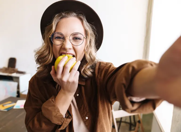 A young blonde woman holds the camera and is taking a large bite of a green apple as takes the photo. She is wearing glasses and a wide-brimmed hat.