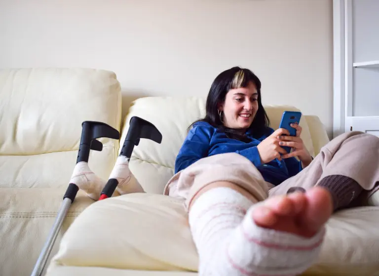 A young white woman with dark hair and a blonde streak sits on her couch. One of her legs is bandaged, and she has it stretched out on a foot stool. A pair of crutches rest against the couch. She’s smiling as she looks at her phone.