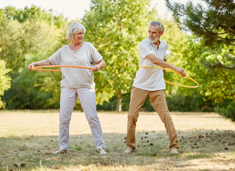 A senior couple stand in a sunny park. Both are holding hula hoops about their waist, ready to start spinning.