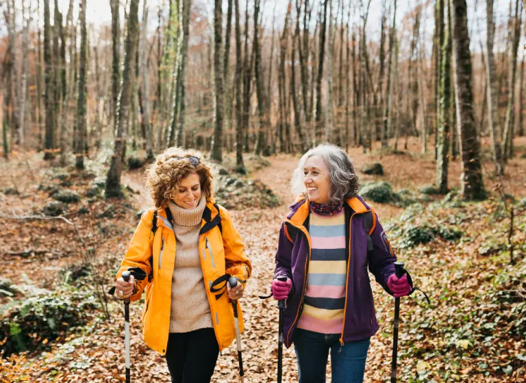 Two mature white women are walking in a forest. They are dressed in warm clothes and the trees have no leaves. Each woman uses poles as they walk.