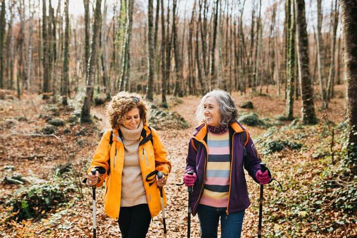 A pair of senior-aged friends use hiking poles as they enjoy a walk in an autumnal forest