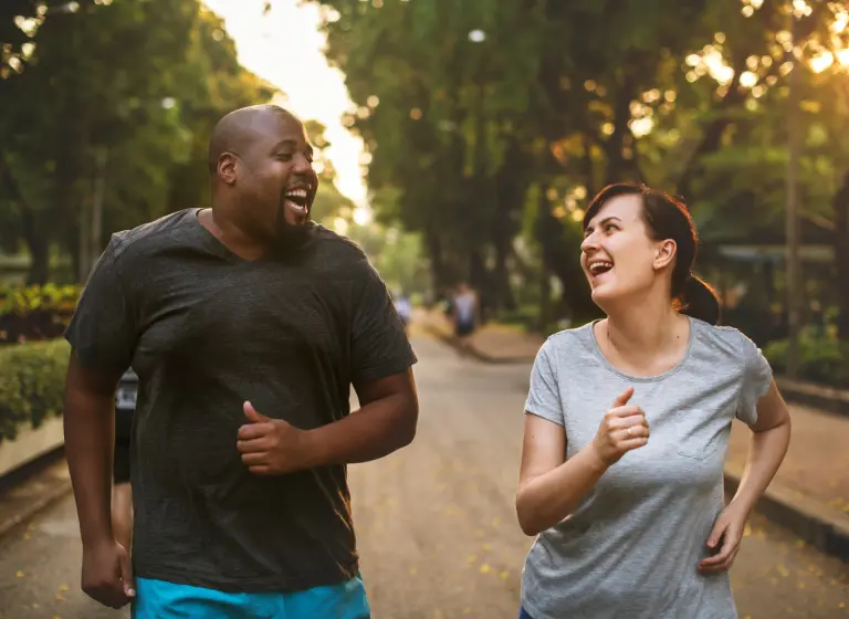 A pair of friends are running along a paved road through a green park as the sun sets. One is a tall, broad black man with a shaved head and neat beard. The other is a shorter white woman with dark hair. They’re looking and smiling at one another.