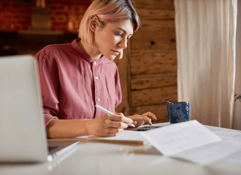 A white woman with jaw-length pastel pink hair sits at her kitchen table with paperwork, her laptop, a calculator, and a mug. She is using the calculator and making notes on a notepad.