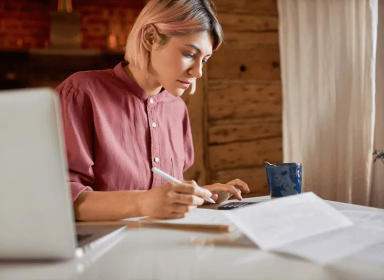A white woman with jaw-length pastel pink hair sits at her kitchen table with paperwork, her laptop, a calculator, and a mug. She is using the calculator and making notes on a notepad. 