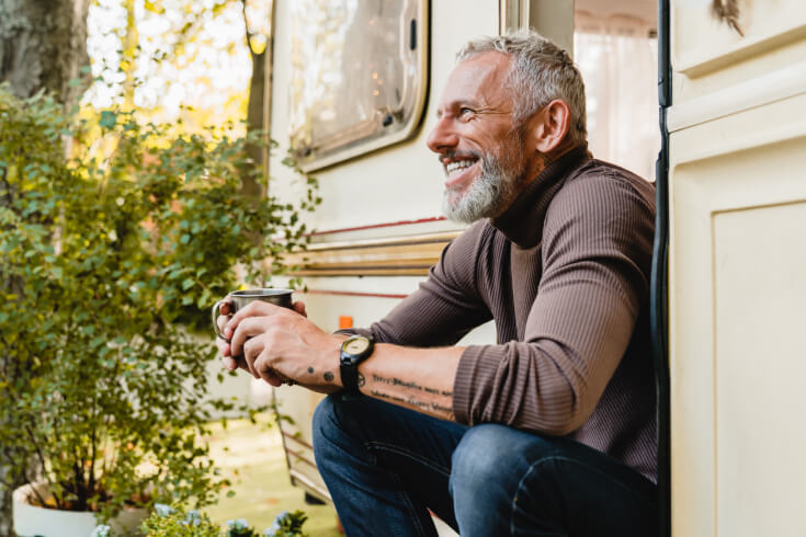 A man in his 60s enjoys a morning coffee while sitting on the step of his caravan