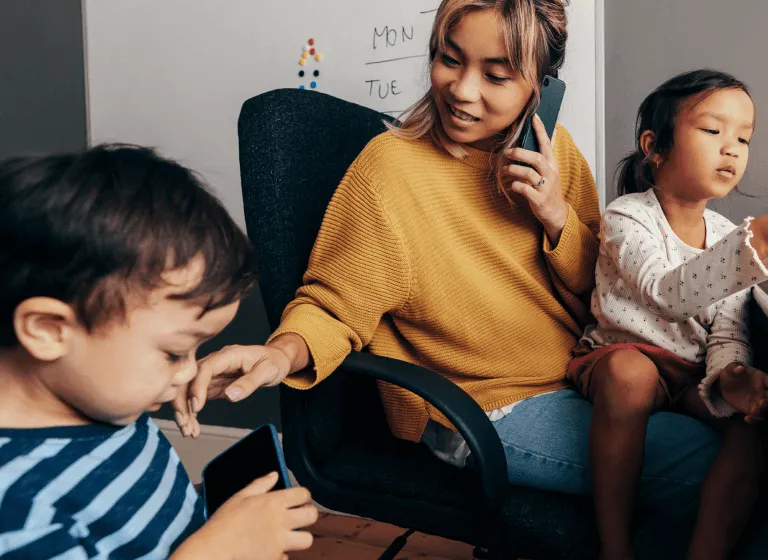 A young Asian woman sits in an office chair with a whiteboard behind her. Her young daughter is sitting on her lap and playing with something off camera. Her young son is standing beside the chair looking at a phone screen. The woman is on her phone while checking what her son is doing.