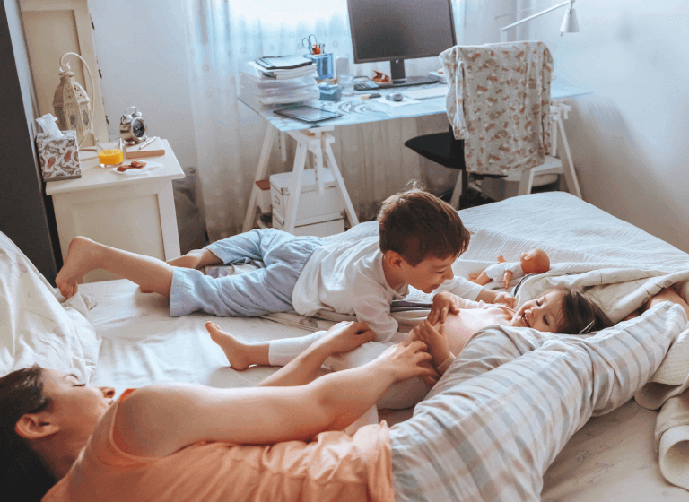 A young mother lies on a double bed with her young son and daughter. They are playing and laughing.