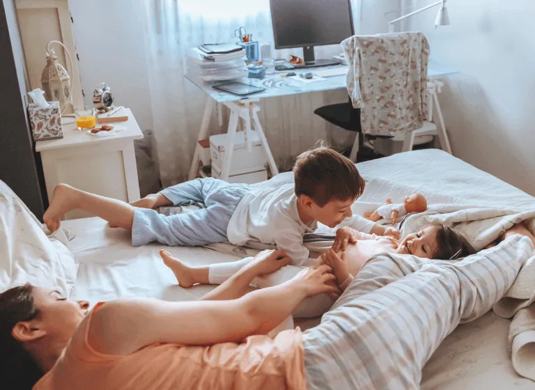 A young mother lies on a double bed with her young son and daughter. They are playing and laughing. 
