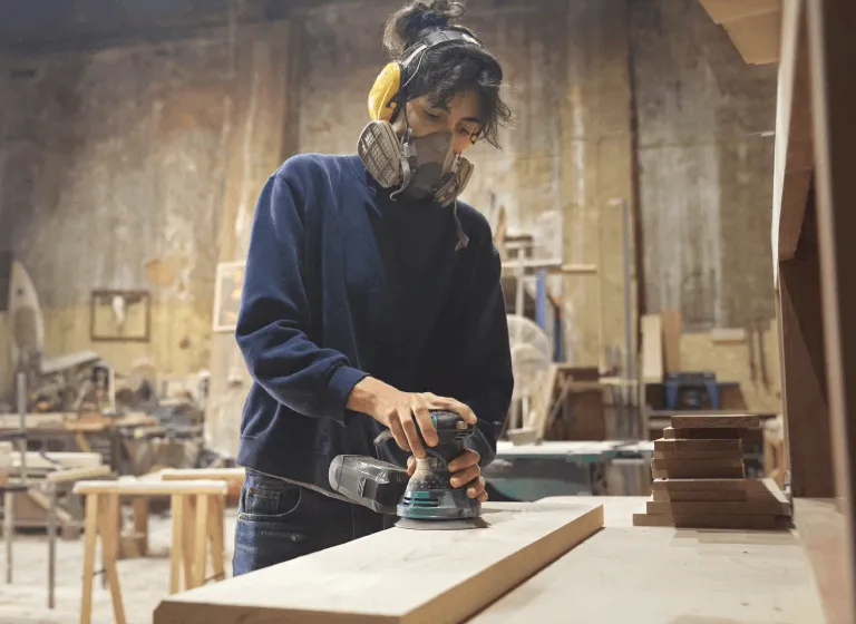 A young white woman is sanding a plank of wood in her workshop. She wears ear proctection and a mask. 