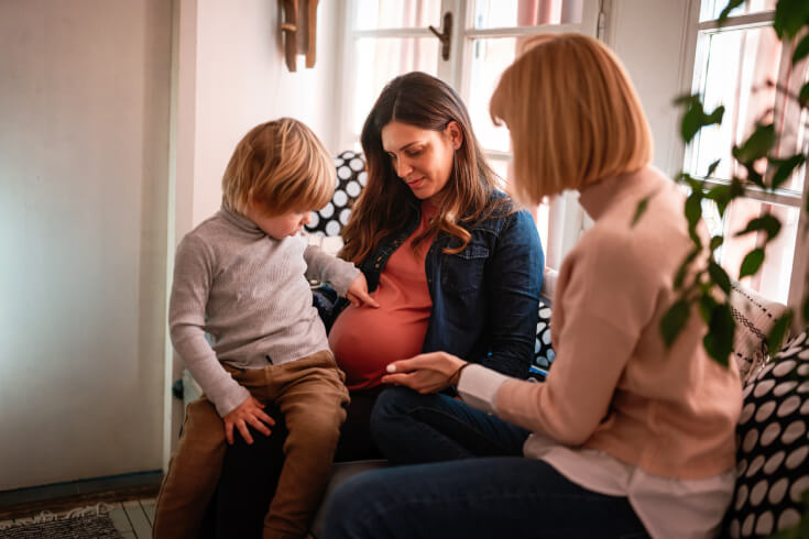 A young child sits on his mother’s lap and gently touches his mother’s pregnant stomach