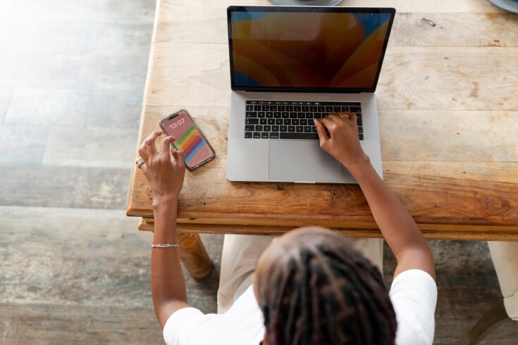 A person types on their laptop while looking at their phone
