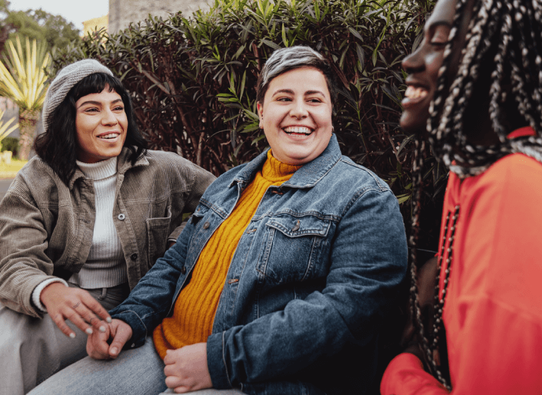 A racially diverse group of young to middle-aged women are sitting outside talking and laughing. 