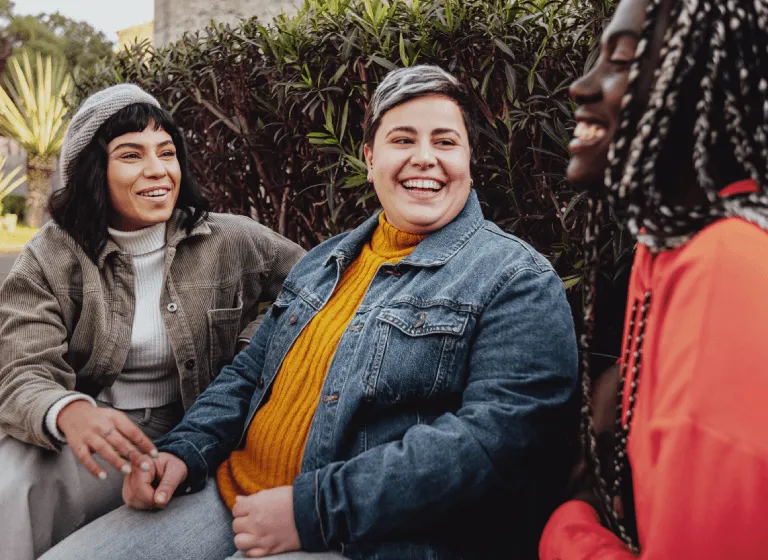A racially diverse group of young to middle-aged women are sitting outside talking and laughing.
