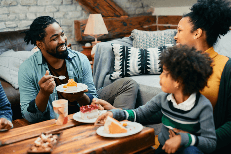 A young black mother and father enjoy cake in their living room with their young child.