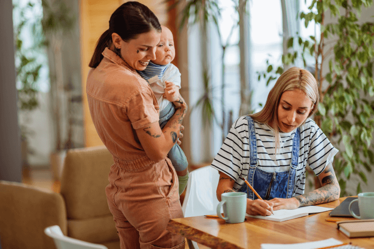 Two young women make notes about health insurance options while one holds a baby