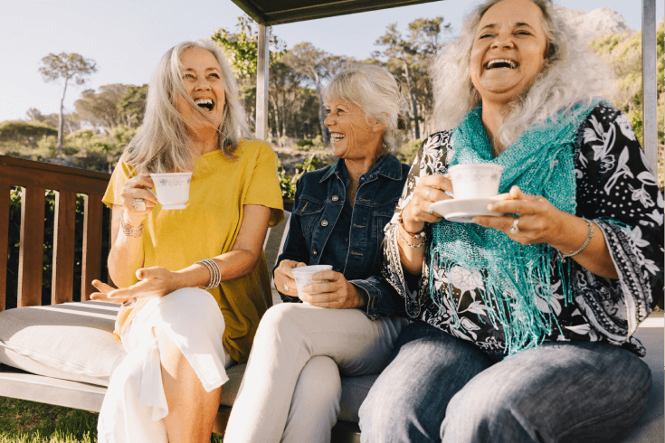 Three laughing older ladies sitting on an outdoor couch, all holding cups of tea.
