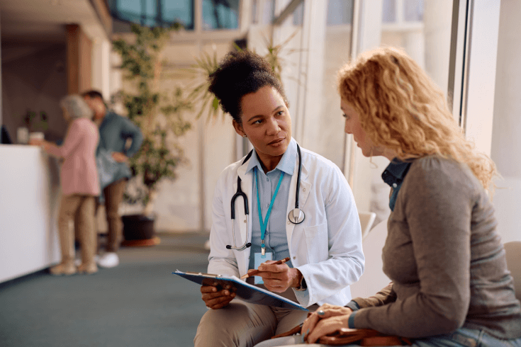 A black female doctor explains a procedure to a white female patient in the waiting room of a hospital.
