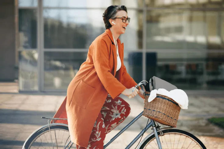 A smiling white woman in an orange coat mounts her bicycle to ride to work, her bag in the basket.
