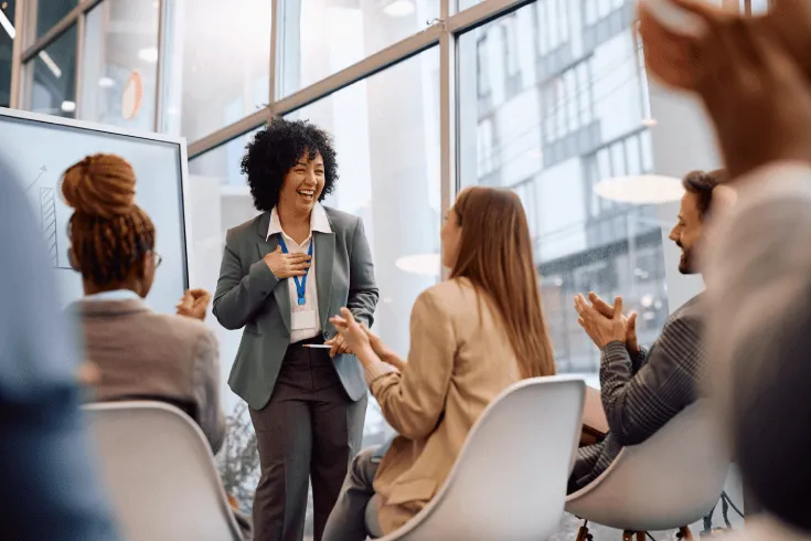A diverse group of colleagues applaud a middle-aged black woman at the end of a business presentation.