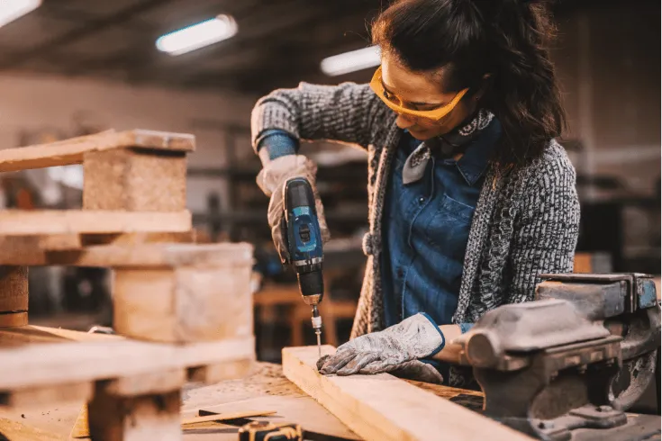 A female carpenter drills into a plank of wood while working in her workshop.