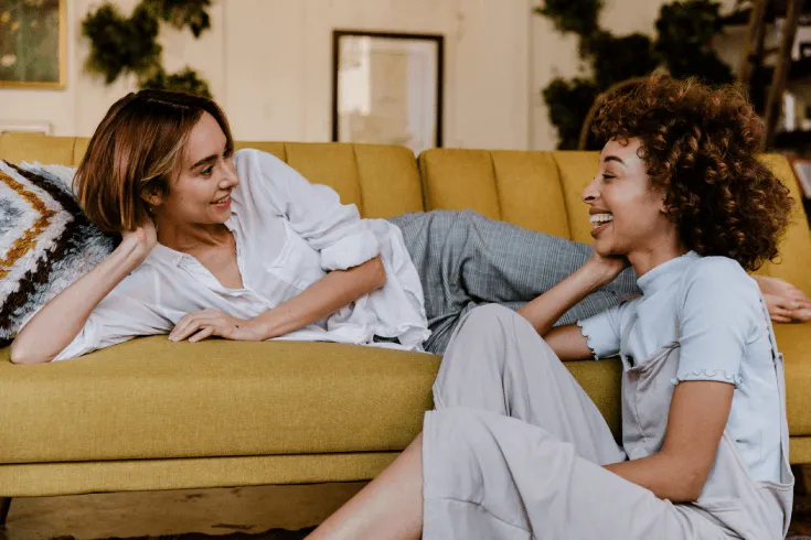A multicultural queer couple laugh as they talk to each and relax at home.