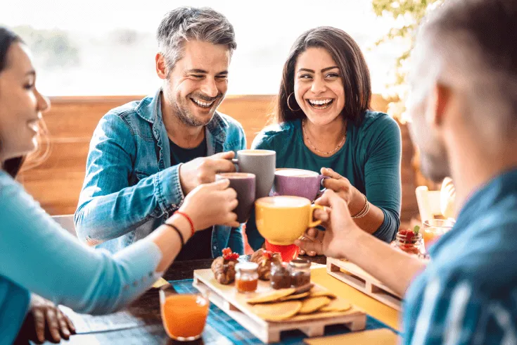 A multicultural group of friends cheers mugs at an outdoor cafe.