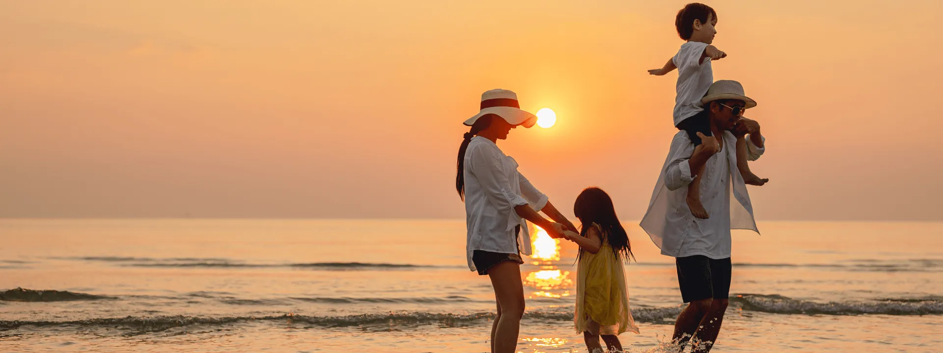 A family stands on a beach at sunset, with the father carrying his son on his shoulders while the mother helps her daughter splash in the waves. 