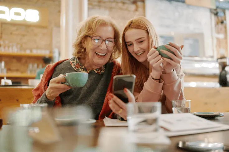 A senior white woman and her daughter look at a phone in a café while holding cups of coffee and smiling.