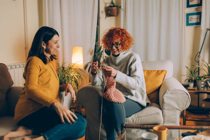 Two middle-aged female friends relax on armchairs as one knits.