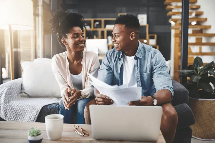 A young black couple smile at each other while sitting on a couch in front of a laptop with paperwork.
