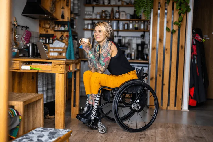 A white woman with tattoos sits in a wheelchair at her kitchen table with a laptop, smiling holding a cup of coffee.