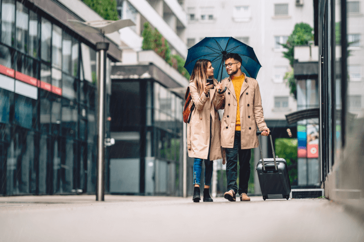 A young couple walks down a rainy street sharing an umbrella and carrying luggage.