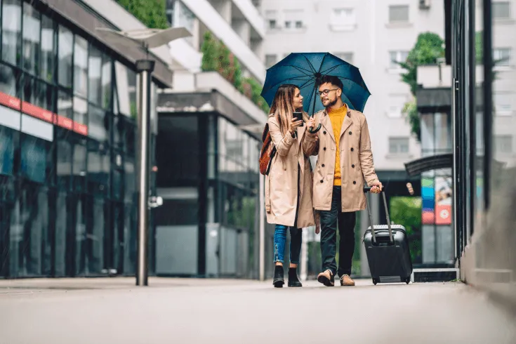 A young couple walks down a rainy street sharing an umbrella and carrying luggage.