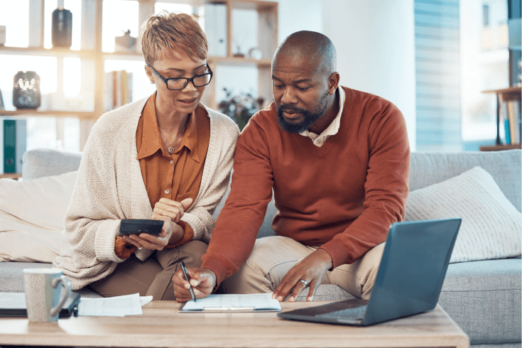 An older black couple sit on a couch, filling out paperwork with the help of a calculator and laptop.