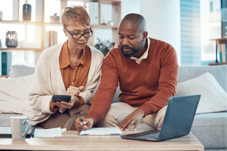 An older black couple sit on a couch, filling out paperwork with the help of a calculator and laptop.