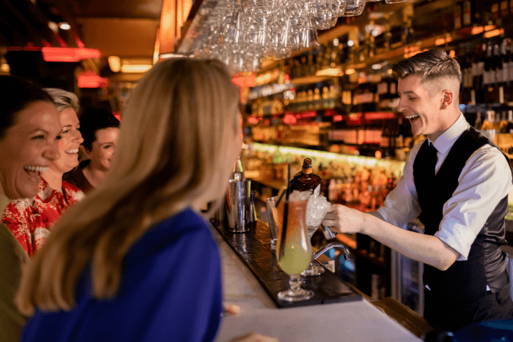 A bartender preparing a cocktail for a group of cheerful women gathered at a bar.