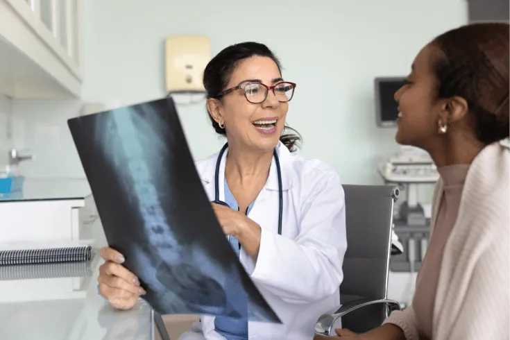 An older olive-skinned female doctor holds up a scan of a woman’s lower pack and hips. Her patient is a young black woman. The doctor is smiling as she explains the scan to her patient. 