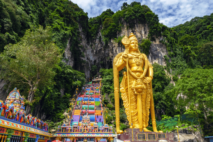 Batu Caves with a golden Hindu statue, rainbow stairs, and tall limestone cliffs in view.