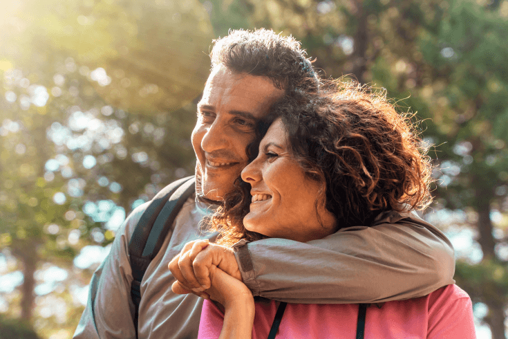 Smiling couple outdoors, with the man wrapping his arm around the woman as they stand close together.