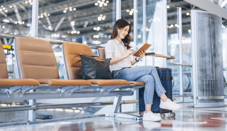 Woman sitting at airport with suitcase and handbag, looking at tablet.