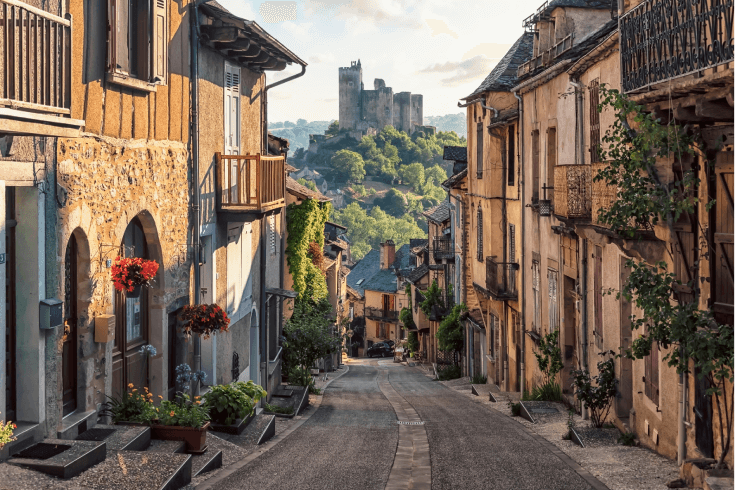 French village street, with buildings on either side and a view of castle ruins on a leafy hill in the distance